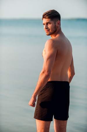 Feeling confident about his shape. Rear view of young muscular man looking at camera over shoulder while standing at the seasideの写真素材