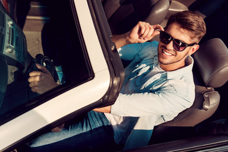 I love this car! Top view of handsome young man looking at camera and adjusting eyewear while driving his white convertibleの写真素材