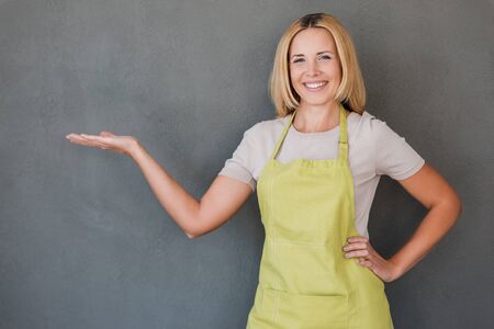 Copy space on her hand. Happy mature woman in green apron holding copy space and smiling while standing against grey backgroundの写真素材