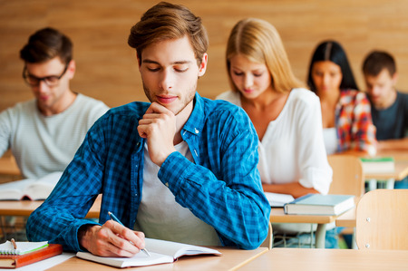 Focused on exam. Group of concentrated young students writing something in their note pads while sitting at their desks in the classroomの写真素材