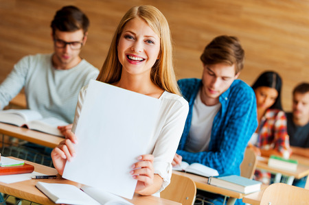 Happy to be the best. Cheerful young woman holding white paper and looking at camera while sitting at front desk in the classroomの写真素材