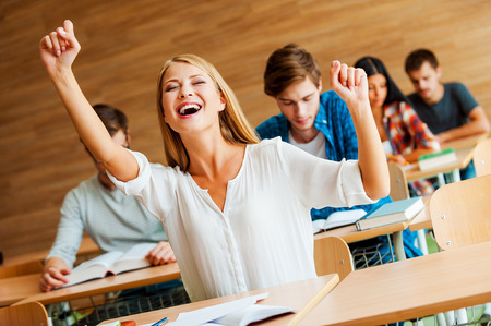 I did it! Excited young woman keeping arms raised while sitting at front desk in the classroomの写真素材