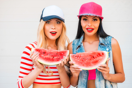 Fruity fun. Two surprised young women holding slices of watermelon and staring at camera while standing outdoorsの写真素材