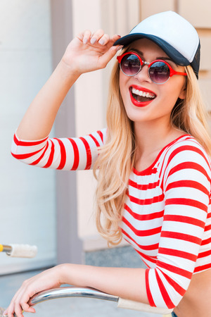 The fun never ends. Joyful young woman adjusting her cap and looking at camera while standing near her bicycle outdoorsの写真素材
