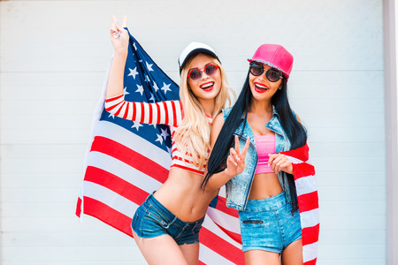 American girls. Two playful young women gesturing peace sign and holding American flag while standing against the garage doorの写真素材