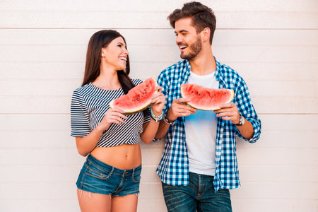 Watermelon lovers. Happy young loving couple holding slices of watermelon and looking at each other while standing outdoorsの写真素材