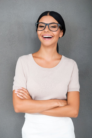 With smile through a life. Cheerful young businesswoman keeping arms crossed and looking at camera while standing against grey backgroundの写真素材