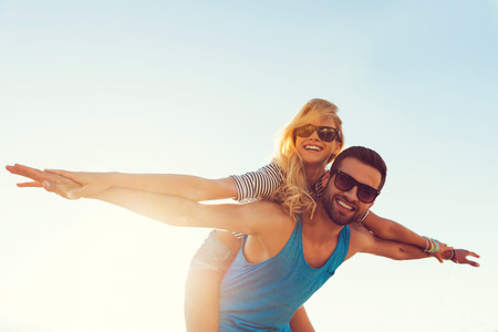 High flying romance. Low angle view of smiling young man piggybacking his girlfriend while keeping arms outstretched High flying romance. Low angle view of smiling young man piggybacking his girlfriend while keeping arms outstretched