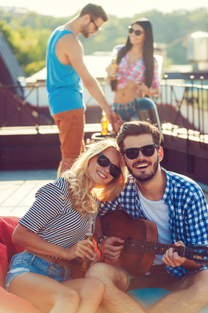 Enjoying time with friends. Beautiful young couple bonding to each other and sitting on the bean bag with guitar while two people barbecuing in the backgroundの写真素材