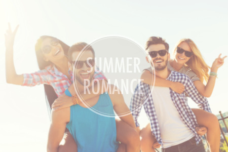 Summer romance. Low angle view of two joyful young men carrying their girlfriends on the shoulders while having fun on the roofの写真素材