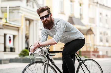 Time to hit the road. Handsome young bearded man looking at camera while sitting on his bicycle outdoorsの写真素材