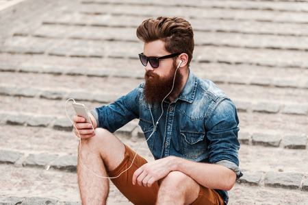 On the city wave. Top view of young bearded man in headphones holding mobile phone while sitting outdoorsの写真素材