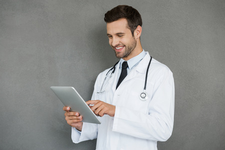Digital solution for medical science. Smiling young doctor in white uniform working on digital tablet while standing against grey backgroundの写真素材