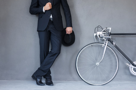 Elegant style. Close-up of young businessmanholding hat and adjusting his jacket while standing near his bicycle against grey backgroundの写真素材