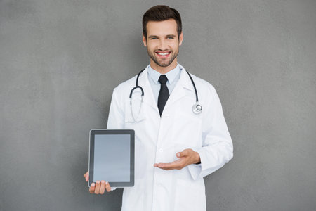 Copy space on his tablet. Cheerful young doctor in white uniform holding digital tablet and pointing at it while standing against grey backgroundの写真素材