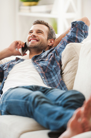 Lazy day at home. Joyful young man talking on the phone while lying on sofaの写真素材