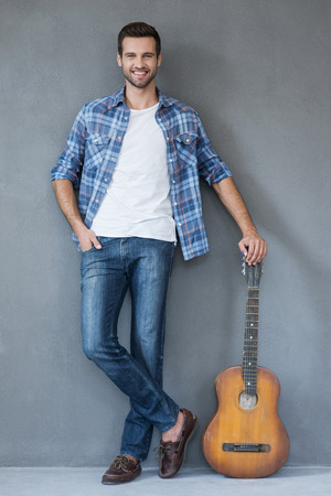My guitar is always with me. Full lengthof young man leaning at the guitar and looking at camera while standing against grey backgroundの写真素材