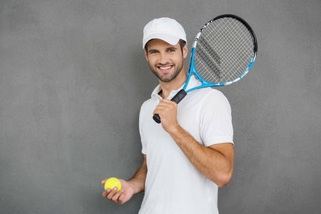 This game is more than hobby! Happy young man in sports clothes carrying tennis racket on his shoulder and smiling while standing against grey backgroundの写真素材