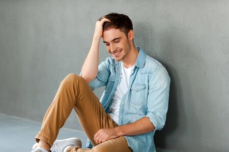 Feeling relaxed. Happy young man holding hand in hair and smiling while sitting against grey backgroundの写真素材