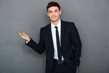 Pointing copy space. Happy young man in formalwear pointing away and looking at camera while standing against grey backgroundの写真素材