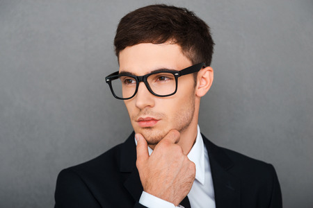 Serious about his business. Confident young businessman holding hand on chin and looking away while standing against grey backgroundの写真素材