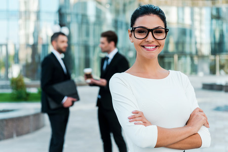 Confident businesswoman. Smiling young businesswoman keeping arms crossed and looking at camera while two her male colleagues talking to each other in the background Confident businesswoman. Smiling young businesswoman keeping arms crossed and looking at camera while two her male colleagues talking to each other in the background