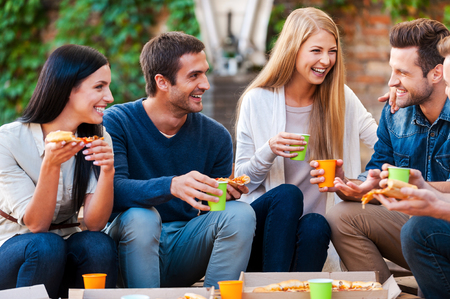 Spending good time with friends. Group of cheerful young people talking to each other and eating pizza while sitting outdoors