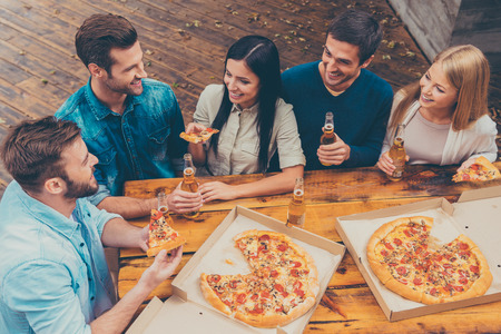 Enjoying time together. Top view of five happy young people holding bottles with beer and eating pizza while standing outdoors