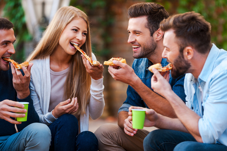 So tasty! Group of joyful young people smiling and eating pizza while sitting outdoorsの写真素材