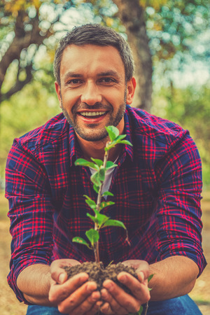 Save the environment! Happy young man stretching out plant in the ground and looking at camera while standing in the gardenの写真素材