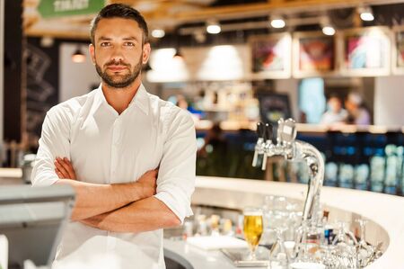 I love my job! Confident young bartender keeping arms crossed and looking at camera while standing at the bar counterの写真素材