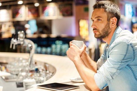 Coffee for inspiration. Side view of young man holding cup of coffee while sitting at the bar...