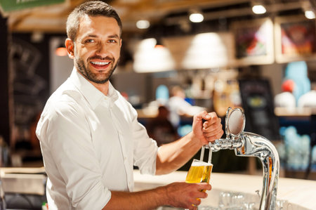 Only fresh beer in his bar! Happy young bartender pouring beer while standing behind bar counter