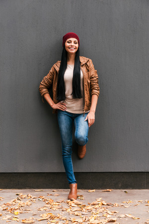 Beauty in style. Beautiful young woman leaning at the grey wall and smiling with fallen leaves laying around herの写真素材