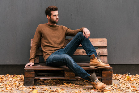 Casually handsome. Handsome young man sitting on the wooden pallet and looking away with grey wall in the background and orange fallen leaves on the floorの写真素材