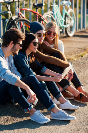 Young and carefree. Close-up of group of young smiling people bonding to each other and looking at digital tablet while sitting outdoors together with bicycles in the backgroundの写真素材