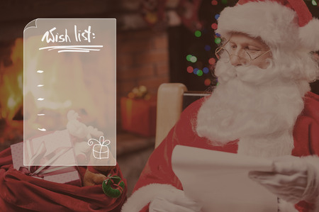 Wish list. Traditional Santa Claus looking at his sack with presents and holding a paper while sitting at his chair with fireplace and Christmas Tree in the backgroundの写真素材