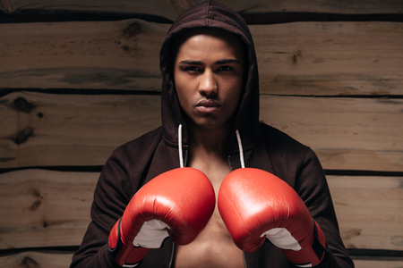 Ready to fight. Confident young African man in hooded shirt and boxing gloves looking at camera while standing against wooden backgroundの写真素材
