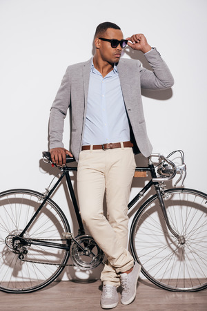 Confident in his perfect style. Full length of confident young African man adjusting his sunglasses and leaning at his retro styled bicycle while standing against white backgroundの写真素材