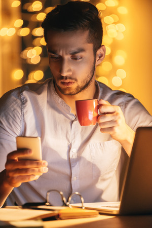 Having no time for break. Confident young man looking at his smart phone and holding coffee cup while sitting at his working place at night time with Christmas lights in the backgroundの写真素材