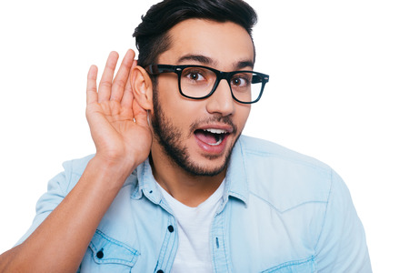 What did you say? Curious young Indian man holing hand near ear and looking at camera while standing against white backgroundの写真素材