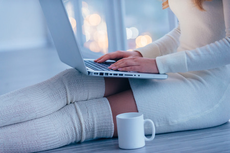 Beauty with laptop. Close-up of beautiful young woman in white sweater and socks working on laptop while sitting on the floor at homeの写真素材