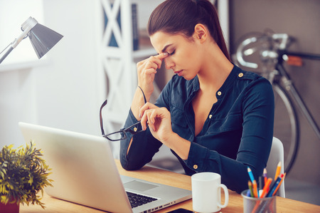 Feeling tired and stressed. Frustrated young woman keeping eyes closed and massaging nose while sitting at her working place in officeの写真素材