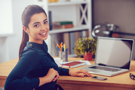 Enjoying her working day. Attractive young woman looking over shoulder and smiling while sitting at her working place in office Enjoying her working day. Attractive young woman looking over shoulder and smiling while sitting at her working place in office