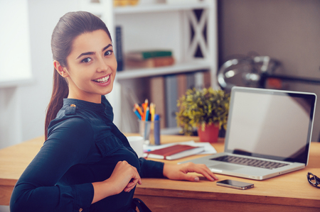 Enjoying her working day. Attractive young woman looking over shoulder and smiling while sitting at her working place in office