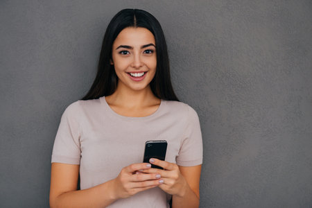 Beauty with smart phone. Attractive young woman holding smart phone and smiling while standing...