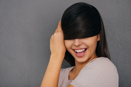 Feeling playful. Beautiful young woman covering eyes with her hair and smiling while standing against grey backgroundの写真素材