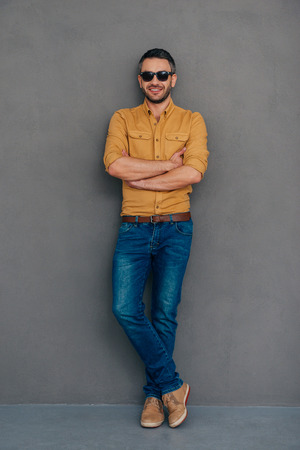 Casually handsome. Full length of confident mature man in sunglasses keeping arms crossed and looking at camera with smile while standing against grey backgroundの写真素材