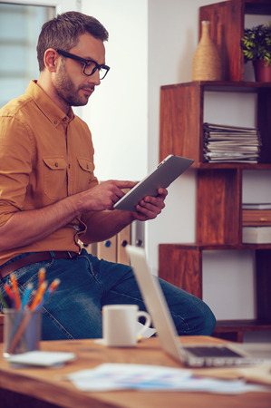 His digital helper. Concentrated young man working on digital tablet while standing near his working place in officeの写真素材