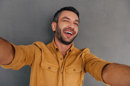 Selfie fun. Smiling young man making selfie and grimacing while standing against grey backgroundの写真素材